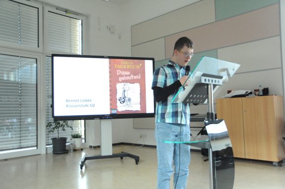 Person steht an einem Stehpult vor einem Bildschirm und präsentiert Informationen.