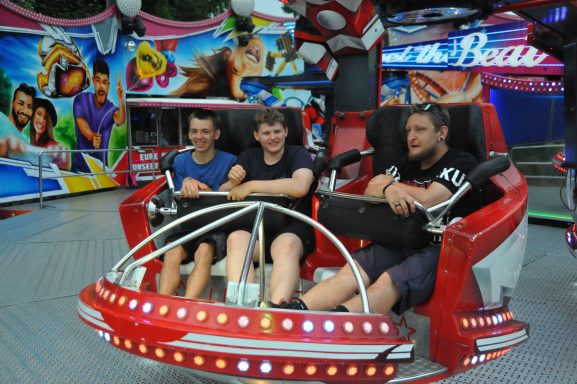Drei Jugendliche sitzen in einer Fahrgeschäft, umgeben von bunten Attraktionen.