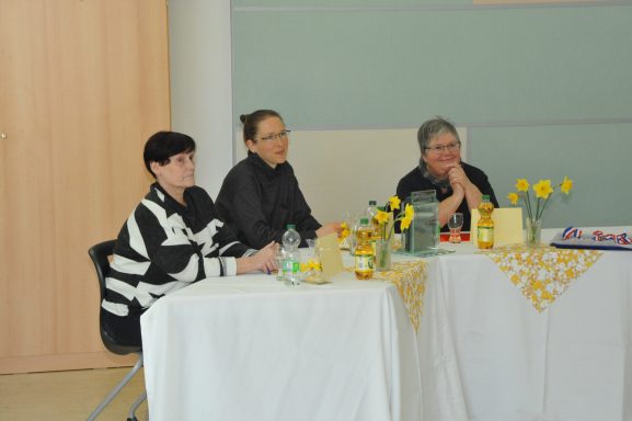 Drei Frauen sitzen an einem Tisch mit Blumen und Getränken, in einem hellen Raum.