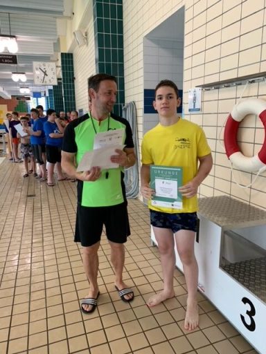 Zwei schwimmende Personen in einem Schwimmbad, einer mit Urkunde in der Hand.
