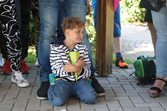 Ein Junge sitzt auf dem Boden, umarmt eine gelbe Tasse, während er von Erwachsenen umgeben ist.