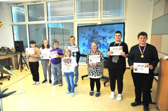 Gruppe von Personen hält Urkunden in einem hellen Raum mit Präsentation im Hintergrund.