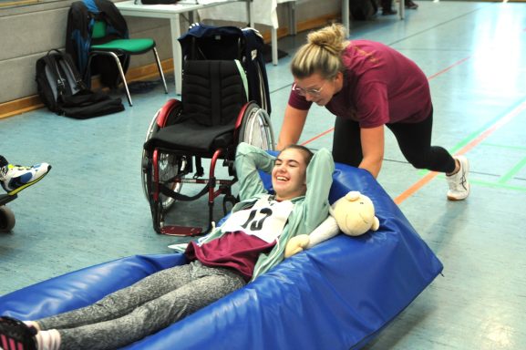 Eine junge Frau liegt auf einer weichen Fläche, während eine zweite Person sie anschiebt.