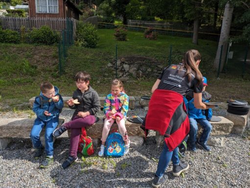 Fünf Kinder sitzen auf einer Bank, während eine Person ihnen etwas reicht.
