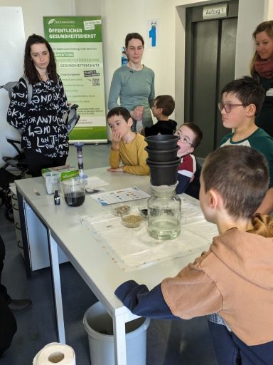 Kinder und Erwachsene bei einem Experiment an einem Tisch in einem Raum mit Aufklärungsposter.