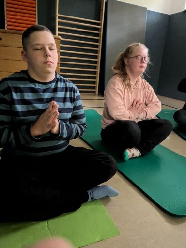 Zwei Personen in Meditationshaltung auf Yogamatten, in einem Raum mit Turngeräten.
