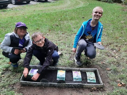Drei Kinder pflanzen Blumen in einem Beet im Freien und lächeln dabei.