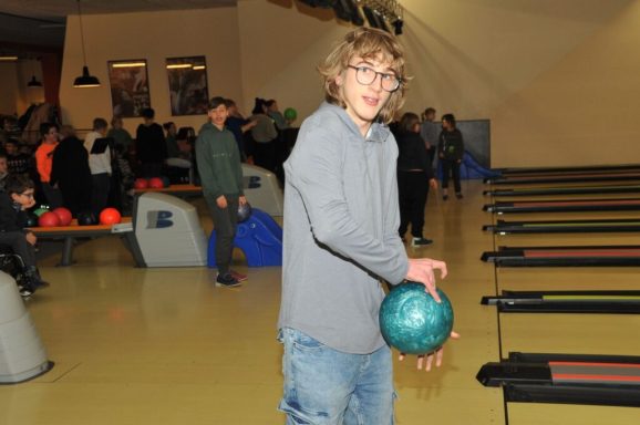Junge mit braunen Locken hält eine Bowlingkugel in einer Bowlingbahn.