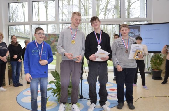 Vier Jugendliche stehen auf einer Bühne mit Medaillen und Urkunden.