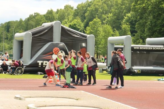 Gruppenspielende Kinder auf einem Sportplatz mit aufblasbaren Hindernissen im Hintergrund.