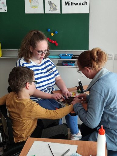 Ein Mädchen und eine Frau helfen einem Jungen beim Basteln im Klassenzimmer.