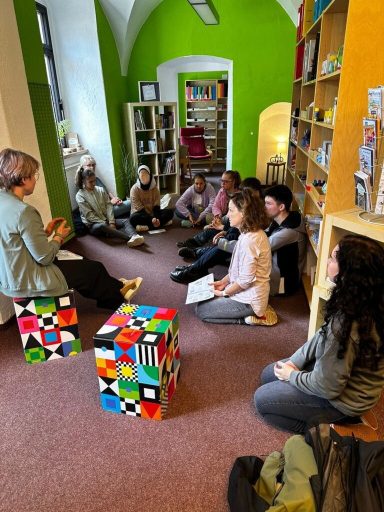 Gruppe von Personen sitzt im Kreis, diskutiert in einem Raum mit bunten Regalen.