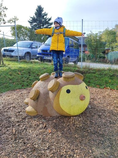 Kind in gelber Jacke balanciert auf einem Igel-Spielgerät auf einem Spielplatz.