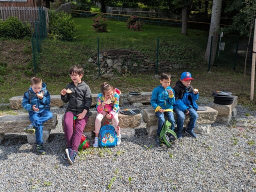 Fünf Kinder sitzen auf einer Steinmauer und essen Snacks in einer grünen Umgebung.