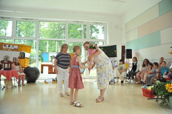 Zwei Frauen überreichen einem Mädchen einen Blumenstrauß in einem festlichen Raum.
