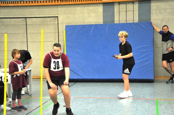 Zwei Personen in Sportkleidung, eine beobachtet, der andere springt über einen Hürdenlauf.