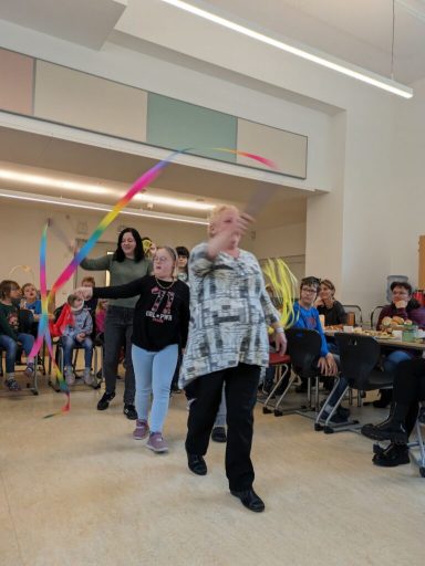 Eine Gruppe von Personen tanzt mit bunten Bändern in einem Raum voller Zuschauer.