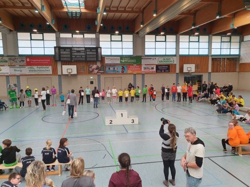 Gruppenvorstellung in einer Sporthalle mit Sportlern in bunten Shirts und Zuschauern.