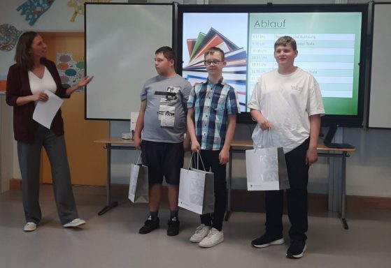 Drei Schüler stehen mit Preisen vor einer Lehrerin und Präsentationsfolien im Hintergrund.