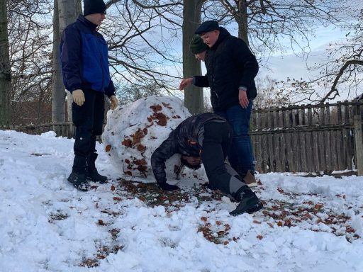 Drei Personen bauen einen großen Schneemann auf einem schneebedeckten Hang.