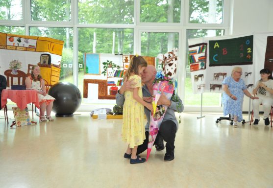 Kinder umarmen sich auf einer Bühne, während andere zuschauen. Bunte Dekoration im Hintergrund.