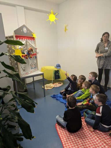 Kinder sitzen auf einer Decke und schauen ein Puppenspiel in einem hellen Raum.