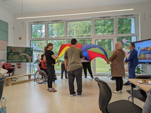 Gruppenspiel mit buntem Fallschirm in einem hellen Raum. Teilnehmer interagieren aktiv.