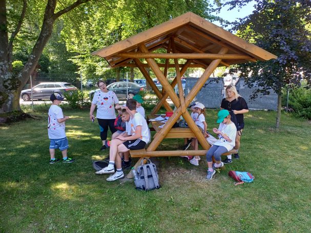 Gruppe von Kindern und Erwachsenen unter einem Holzpavillon im Freien, umgeben von Bäumen.