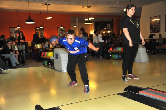 Ein Kind in blauer Uniform wirft einen Bowlingball, während eine Erwachsene zuschaut.