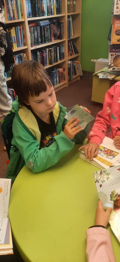 Ein Kind in grüner Jacke hält einen Würfel an einem Tisch in einer Bibliothek.