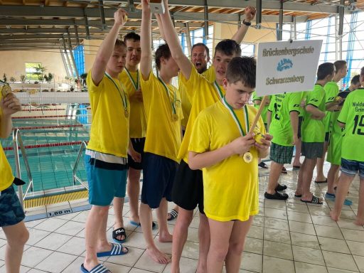 Sechse Jungen in gelben T-Shirts mit Medaillen stehen am Schwimmbecken und halten ein Schild.
