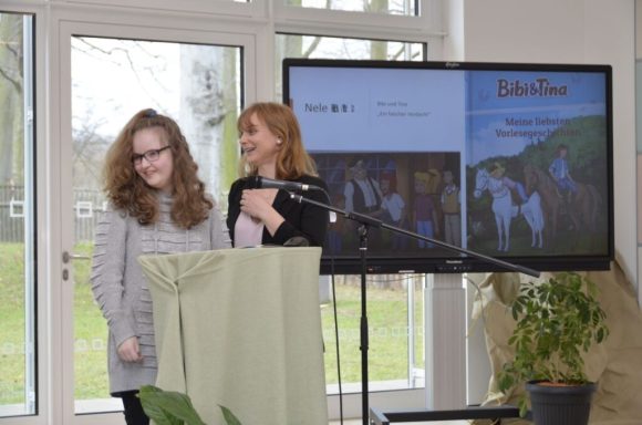 Zwei Frauen stehen neben einem Tisch während einer Präsentation, im Hintergrund ein Bildschirm.