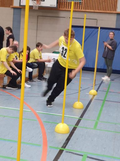 Ein Athlet in gelbem Shirt läuft zwischen aufgestellten Stangen in einer Sporthalle.