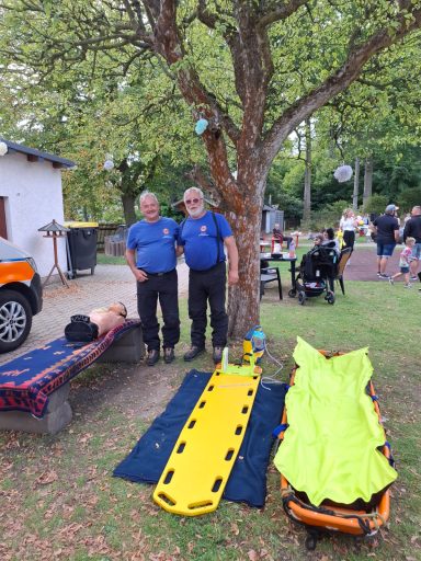 Zwei Männer in blauen Shirts stehen neben einer Baum, Sanitätsmaterial und Fahrzeuge im Hintergrund.