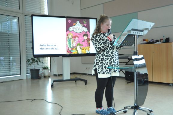 Eine Person spricht an einem Mikrofon vor einer Präsentation mit buntem Bild.