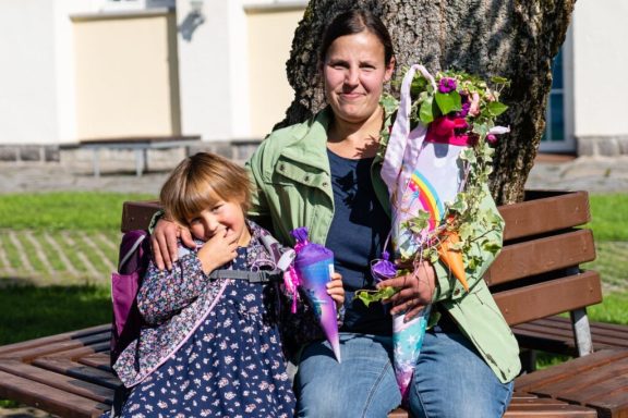 Frau mit Blumenstrauß sitzt auf einer Bank, neben ihr ein lachendes Mädchen.