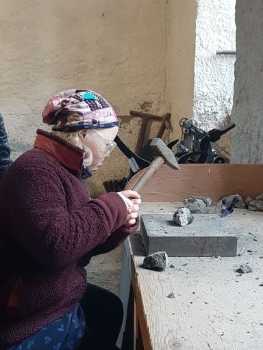 Eine Frau mit Hammer arbeitet an einem Stein in einem handwerklichen Umfeld.