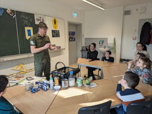 Eine Person steht vor einer Tafel, während Kinder und Erwachsene aufmerksam zuhören.