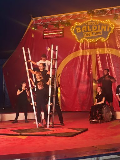 Zirkusaufführung mit Akrobaten auf Leitern vor rotem Vorhang und Publikum im Hintergrund.