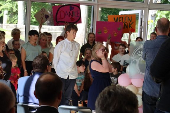 Menschenmenge mit Schildern, einige Zuschauer stehen auf, während ein Redner spricht.