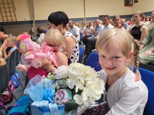 Ein Mädchen mit blonden Haaren hält einen Blumenstrauß und lächelt in einer Menschenmenge.
