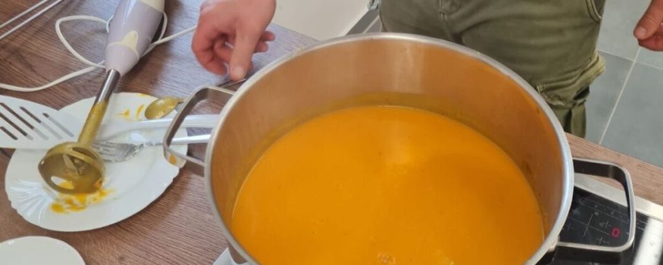Topf mit orangefarbener Suppe und einem Pürierstab auf einem Tisch.