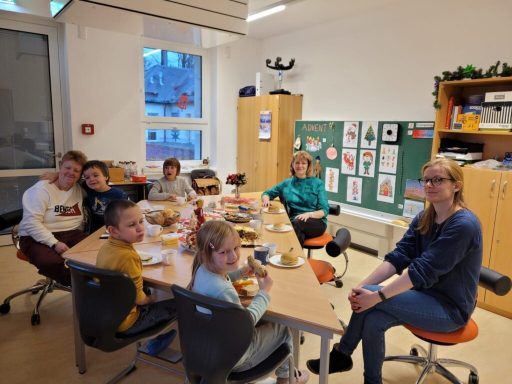 Gruppe von Kindern und Erwachsenen beim Essen an einem Tisch in einem Klassenraum.