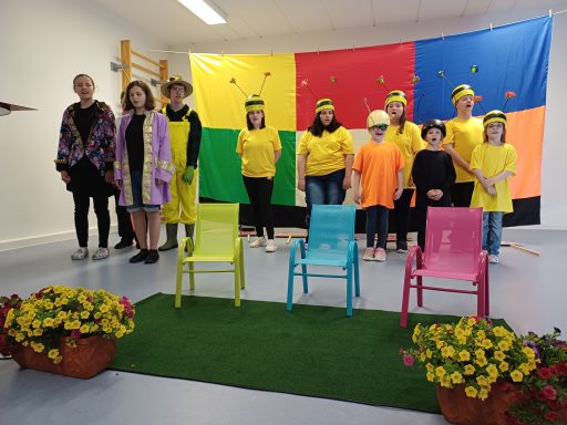 Gruppe von Kindern in bunten Kostümen auf einer Bühne mit Pflanzen im Vordergrund.
