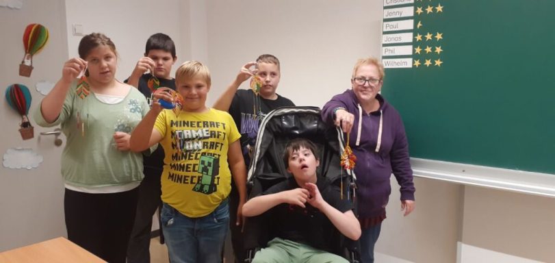 Sechs Kinder und Jugendliche in einem Klassenraum, lächeln und halten Gegenstände in der Hand.