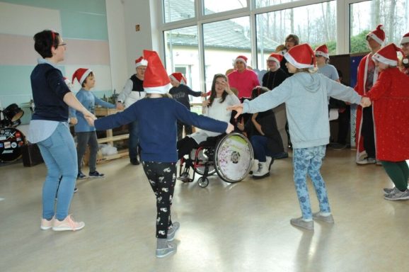 Kinder und Erwachsene mit Weihnachtsmützen tanzen fröhlich in einem Raum.