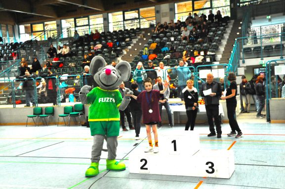 Maskottchen steht mit einem Sieger auf dem Podest in einer Sporthalle. Zuschauer im Hintergrund.