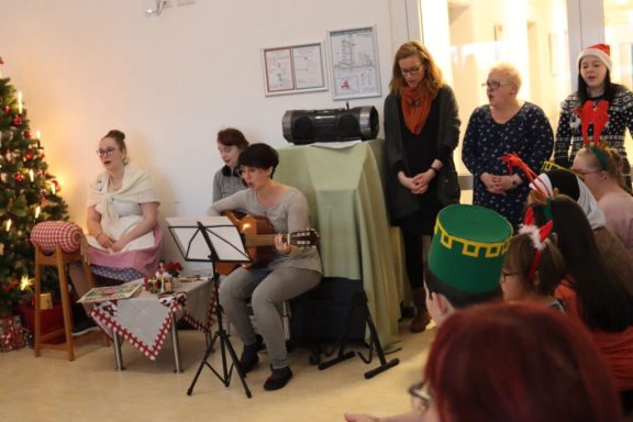 Weihnachtsfeier mit Musikern und Zuschauern, geschmückter Baum im Hintergrund.
