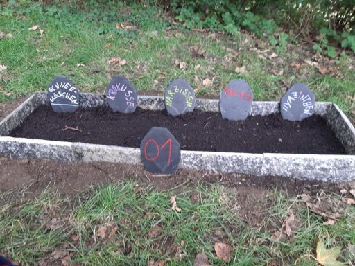 Reihen von kleinen Steintafeln in einem bepflanzten Beet, einige beschriftet.