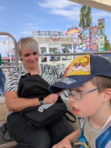 Mutter sitzt hinter einem Jungen auf einer Freizeitveranstaltung, bunte Attraktionen im Hintergrund.